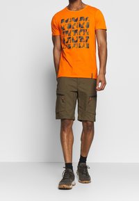 Oranssi puuvillainen t-paita, jossa on musta graafinen kuvio, yhdistettynä oliivinvihreisiin cargo-shortsseihin, joissa on vetoketjulliset taskut ja rento leikkaus.