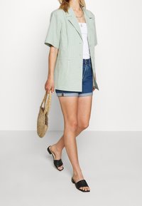 Light green checkered blazer with short sleeves, worn over a white top and denim shorts; paired with black slide sandals and a straw bag.