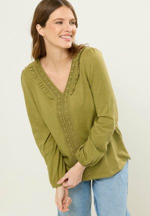 Uśmiechnięta kobieta poprawiająca mankiet oliwkowozielonej bluzki z koronkowym wykończeniem, ubrana w jasnoniebieskie dżinsy, stojąca na tle jasnego tła.