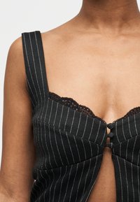 Femme portant un haut sans manches noir à rayures avec bordure en dentelle et trois boutons recouverts de tissu au niveau de la poitrine.