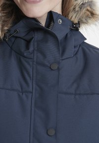 Mörkblå jacka med pälsfodrad huva, som har en hög krage, tryckknappsstängningar och justerbara snoddar för en snug passform.