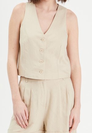 Femme portant un gilet beige sans manches boutonné associé à un short plissé taille haute assorti.