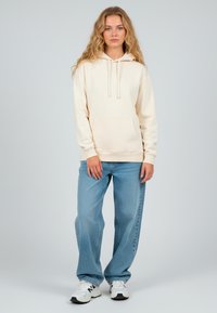 Felpa beige con cappuccio, cordini e tasca frontale, abbinata a jeans larghi di colore blu chiaro. Scarpe da ginnastica bianche completano l'outfit.