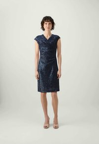 Navy plava haljina sa šljokicama, uskog kroja, kratkim rukavima i body wrap stilom, uparena sa svijetlim štiklama.