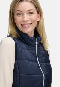 Marineblaues Steppweste mit hohem Kragen, gestepptem Design, silbernem Reißverschluss, getragen über einem hellen Langarmoberteil.