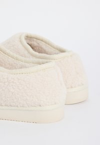Un par de zapatillas de felpa color crema con suelas texturizadas, mostradas desde la parte trasera sobre un fondo blanco.