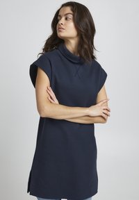 Marineblå ærmeløs tunika med høj cowl-hals, sideslidser og blødt, tekstureret stof. Enkelt design med minimal detaljering.