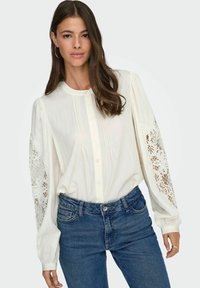 Biała bluzka z guzikami, wykonana z teksturowanej tkaniny, z koronkowymi akcentami na rękawach, zestawiona z niebieskimi dżinsami. Długie, pufiaste rękawy i subtelne plisy.