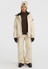 Beige waterdichte jas met een zwarte voering, gecombineerd met beige sneeuwbroek. Zwarte handschoenen en een beige beanie maken de buitenoutfit compleet.