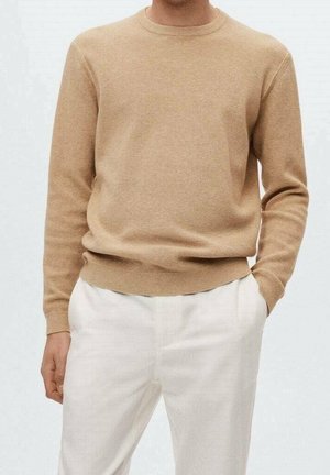 Homme portant un pull en tricot beige à manches longues et un pantalon blanc, une main dans la poche, debout devant un fond uni.