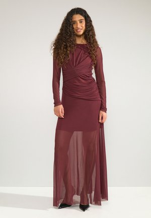 Femme aux longs cheveux bouclés portant un haut froncé bordeaux à manches longues et une jupe longue transparente avec des chaussures noires pointues.