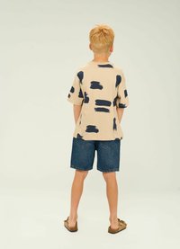 Beige t-shirt med kort ärm och marina penseldragspattern, parat med denimshorts och bruna slip-on-skor. Vy bakifrån.