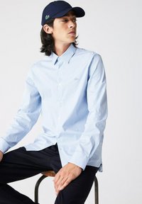 Camicia azzurra a righe con colletto classico, maniche lunghe e un piccolo logo verde sul petto. Indossata con pantaloni scuri.
