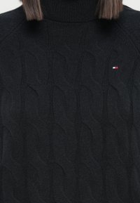 Pullover nero lavorato a maglia con motivo a treccia, con colletto alto. Piccolo logo rosso, bianco e blu sul petto. Tessuto morbido.