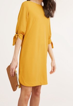 Kvinde i en knælængde kjole i sennepsgul med 3/4-ærmer bundet ved albuerne, der holder en brun flettet clutch.