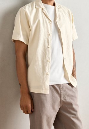 Mann trägt beige kurzärmliges Hemd mit Knöpfen über weißem T-Shirt und hellgrauer Hose, steht vor einer schlichten hellen Wand.