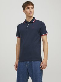 Marineblå polo shirt lavet af bomuld med en tre-knaps placket, hvid-rød-blå stribet krave og lille logo på brystet. Båret med denim jeans.
