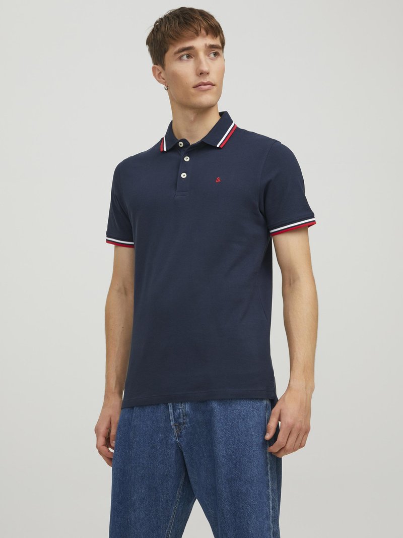 Marineblå polo shirt lavet af bomuld med en tre-knaps placket, hvid-rød-blå stribet krave og lille logo på brystet. Båret med denim jeans.