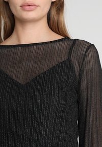Maglietta nera a maniche lunghe con design in rete trasparente, caratterizzata da righe verticali e scollatura a V, arricchita con fili metallici sottili.