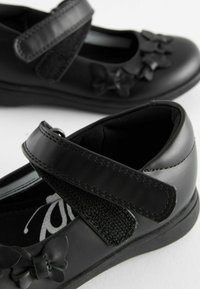 Chaussures en cuir noir avec un bout arrondi, dotées d'une sangle en Velcro et de décorations en forme de papillon noir à l'avant. Semelle texturée.