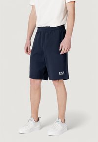 Shorts de sport bleu marine avec taille élastique, longueur genoux, texture lisse, arborant un petit logo EA7 blanc sur la jambe inférieure.