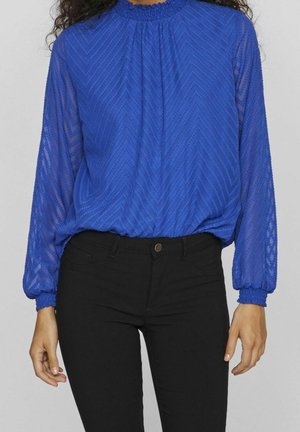 Femme portant un chemisier bleu à motifs avec manches longues et poignets smockés, et un pantalon noir ajusté, debout devant un fond uni.