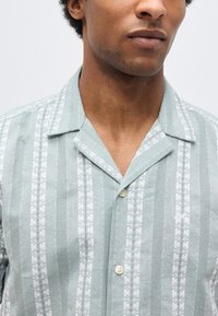 Άνδρας που φοράει ανοιχτό πράσινο πουκάμισο με κάθετες ρίγες από λευκά και πράσινα λουλουδένια σχέδια, με ανοιχτό γιακά και υφασμάτινη υφή.