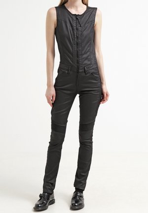 Sorte ærmeløse jumpsuit med en frontlynlås, strukturerede paneler ved knæene og et slankt, tætsiddende design. Bæres med sorte sko.