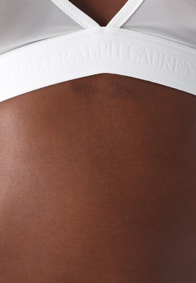 Balts bralete ar acs dizainu un teksturētu joslu, kurā ir izspiestais "POLO RALPH LAUREN" logotips, uz brūna fona.