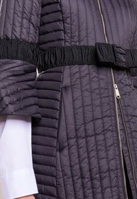 Quiltad lila dunjacka med svart elastiskt midjebälte och sidoficka med dragkedja. Har vida ärmar och en slät, glänsande yta.