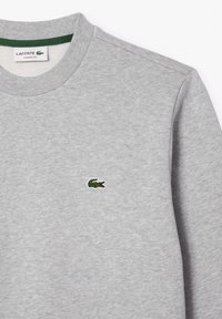 Sweat-shirt gris clair col rond avec petit logo crocodile vert brodé sur la poitrine et étiquette de la marque Lacoste à l’intérieur du col.