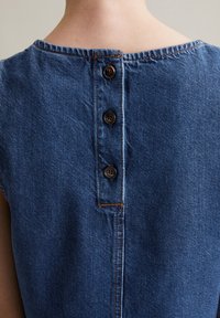 Denimowa bluzka z okrągłym dekoltem, z trzema brązowymi guzikami z tyłu i widocznymi szwami, w średnim do ciemnego niebieskim odcieniu.