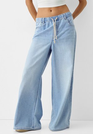 Femme portant un jean bleu clair à jambes larges avec une taille élastique et un cordon blanc, associé à un haut court blanc, debout devant un fond blanc.