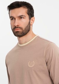 Beige Langarmhemd mit einem hellen, runden Halsausschnitt. Auf der Brust befindet sich ein gesticktes Logo in Beige. Weiche Stofftextur.