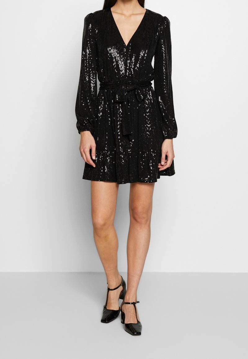 Robe noire en sequins avec un décolleté en V profond, des manches longues bouffantes, une ceinture à la taille et une jupe évasée. Associée à des escarpins noirs en vernis.