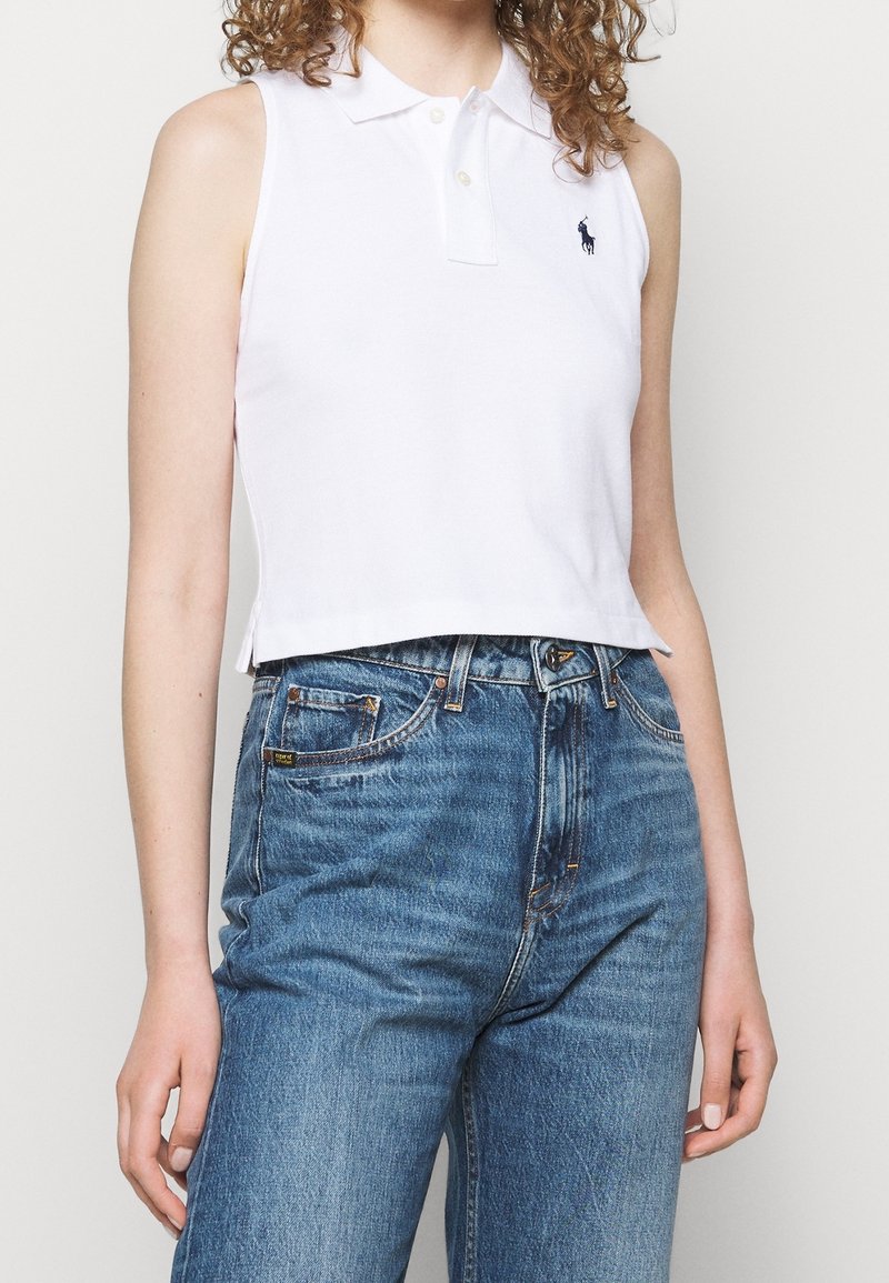 Femme portant un polo blanc sans manches avec un petit logo brodé et un jean bleu taille haute avec coutures et poches visibles.