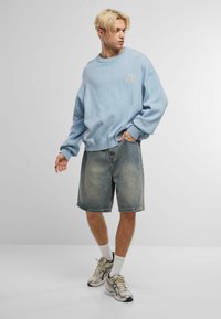 Világoskék, oversized pulóver bordázott mandzsettákkal, világoskék farmer rövidnadrággal és bézs sportcipővel párosítva, diszkrét logó részlettel.
