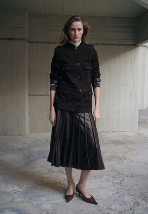 Kobieta stojąca w pomieszczeniu, ubrana w ciemną kurtkę zapinaną na guziki, plisowaną skórzaną spódnicę midi oraz szpilki na betonowej podłodze.