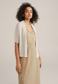 Világoskrém színű, rövid ujjú sál, amely lazán illeszkedik és puha tapintású, arany pliszírozott ruhára viselve, rugalmas derékkal.