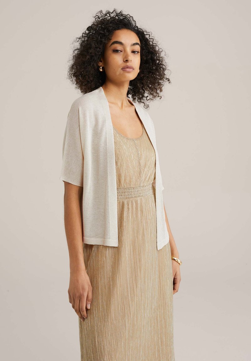 Világoskrém színű, rövid ujjú sál, amely lazán illeszkedik és puha tapintású, arany pliszírozott ruhára viselve, rugalmas derékkal.