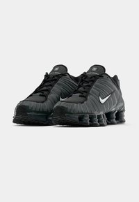Juodų Nike Shox bėgimo batų pora su baltomis swoosh logotipų, turinčių spyruoklinio tipo paminkštinimus, ant baltos fono.