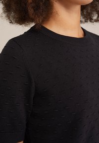 Melna adīta džemperis ar teksturētu rakstu, kurā ir mazi pacelti punkti, ribināta apkakle un īsas piedurknes.
