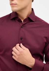 Bordeauxkleurig shirt van een gestructureerde stof, met een button-down kraag en zwarte knopen. Een hand is zichtbaar terwijl deze de voorkant van de sluiting aanpast.