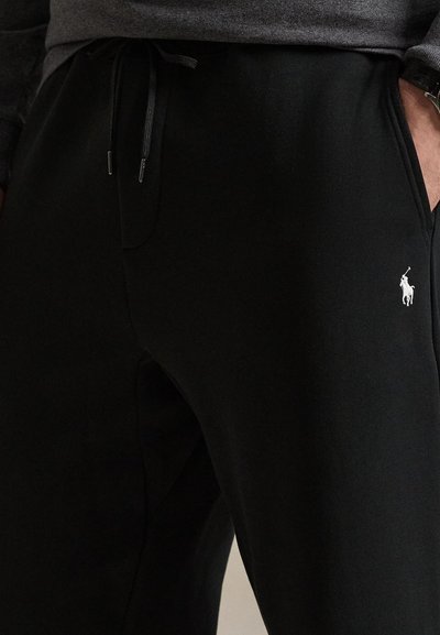 Close-up af en person iført sorte joggingbukser med snøre og en lille hvid Polo-logo nær den højre lomme.
