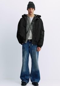 Schwarze Bomberjacke mit Kapuze, graues kariertes Hemd darunter, weißes T-Shirt, blaue Schlagjeans, schwarze Stiefel und dunkle Strickmütze.