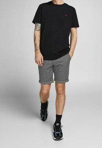 Mężczyzna w czarnej koszulce, szarych szortach zwiniętych do góry, czarnych skarpetach i czarnych sneakersach, idzie z jedną ręką w kieszeni, widoczną tatuażową ręką.