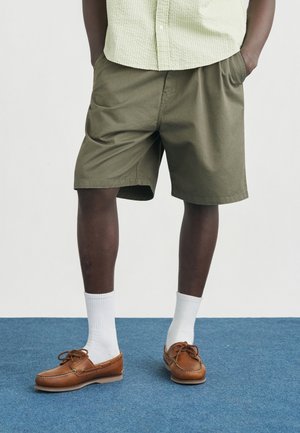 Olívazöld térdig érő rövidnadrágot, fehér zoknit és barna bőrcsónakcipőt viselő férfi kék szőnyegen áll.