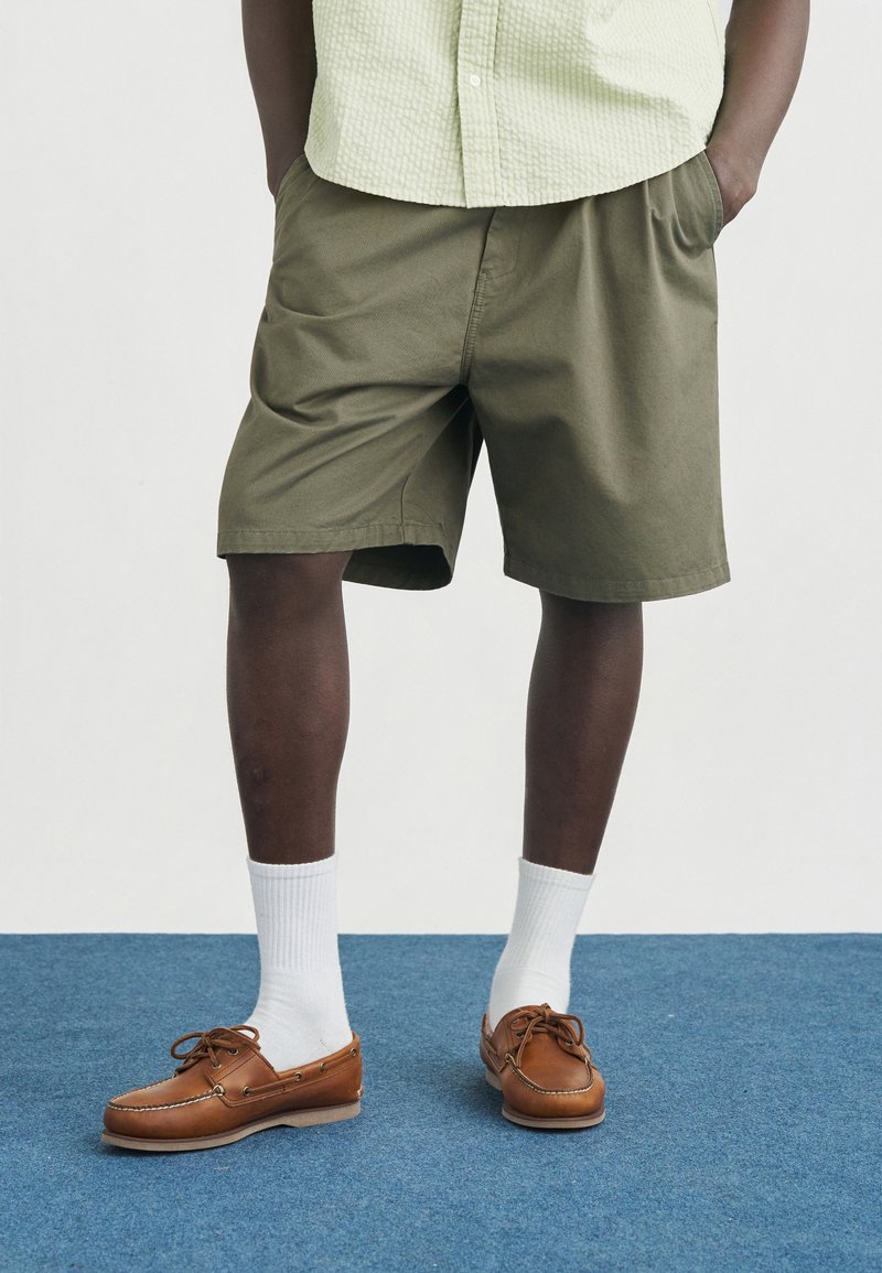 Άνδρας που φοράει χακί βερμούδα μέχρι το γόνατο, λευκές κάλτσες μέχρι τον αστράγαλο και καφέ δερμάτινα παπούτσια για καμπίνα, στέκεται σε μπλε χαλί.