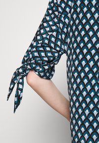Robe imprimée présentant un motif de vagues noir, teal et bleu clair. La manche est nouée avec des accents en tissu au niveau du coude. Texture soyeuse.