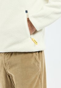 Pull en polaire crème avec une poche zippée, mettant en avant un accent bleu marine à l'ouverture ; associé à un pantalon en velours côtelé beige.